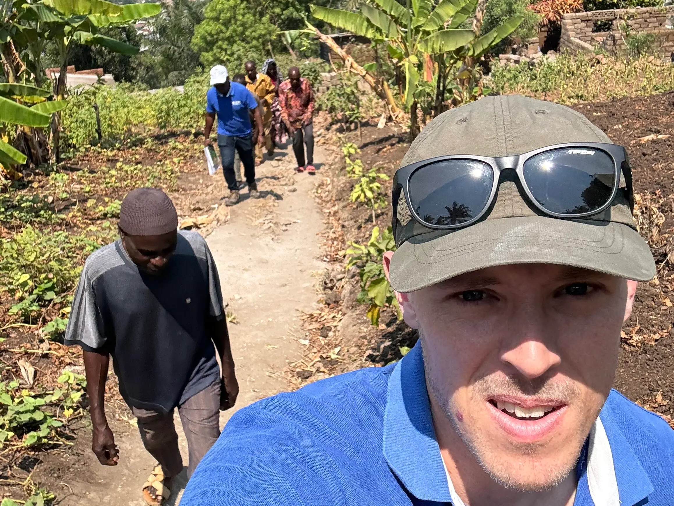Person in a blue UNHCR/IOM shirt and sunglasses takes a selfie on a rural trail with others walking behind, surrounded by lush greenery and a view of a coastal town in the distance.