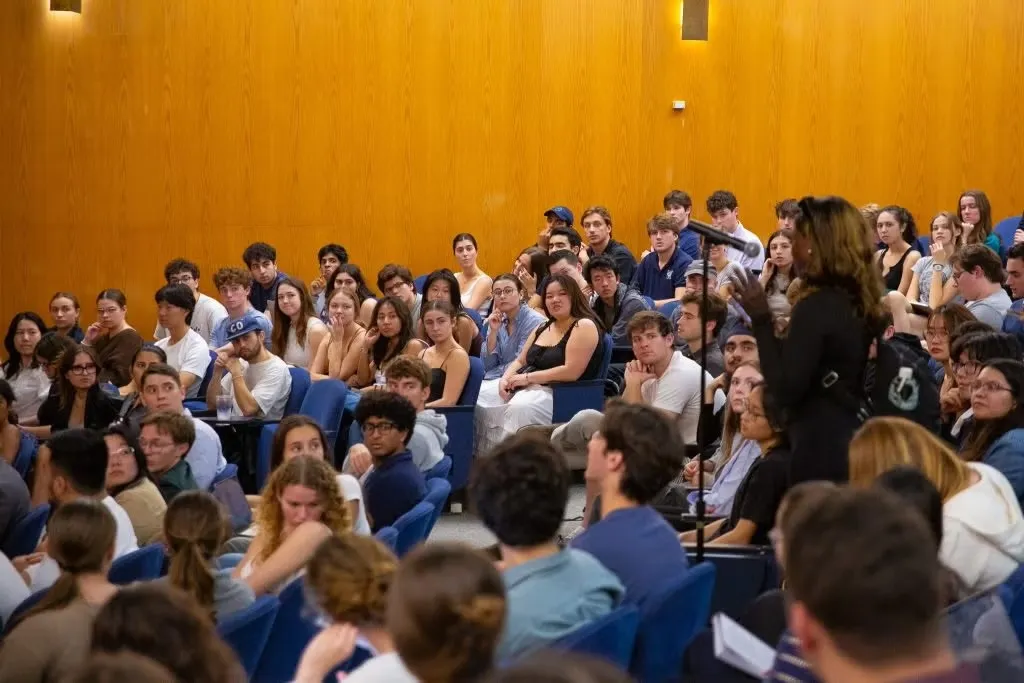 Audience of students attentively sitting in a lecture hall. A woman with long brown hair wearing a black dress asks a question at the microphone.