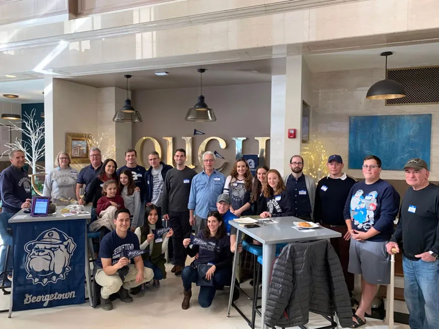 Group of Georgetown alumni and their families gathered at an indoors alumni event, all wearing navy blue or gray shirts, holding Hoya Saxa flags.