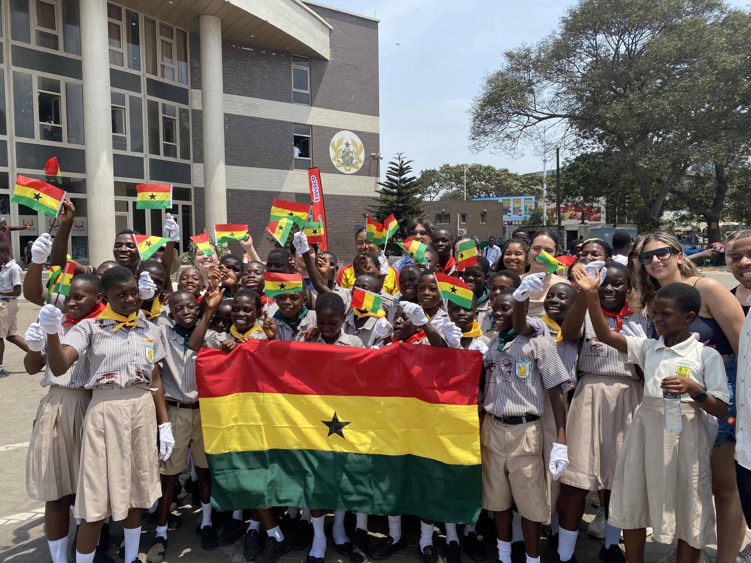 Large group of Ghanian children in school uniforms hold large flag of Ghana while waving small flags of Ghana.