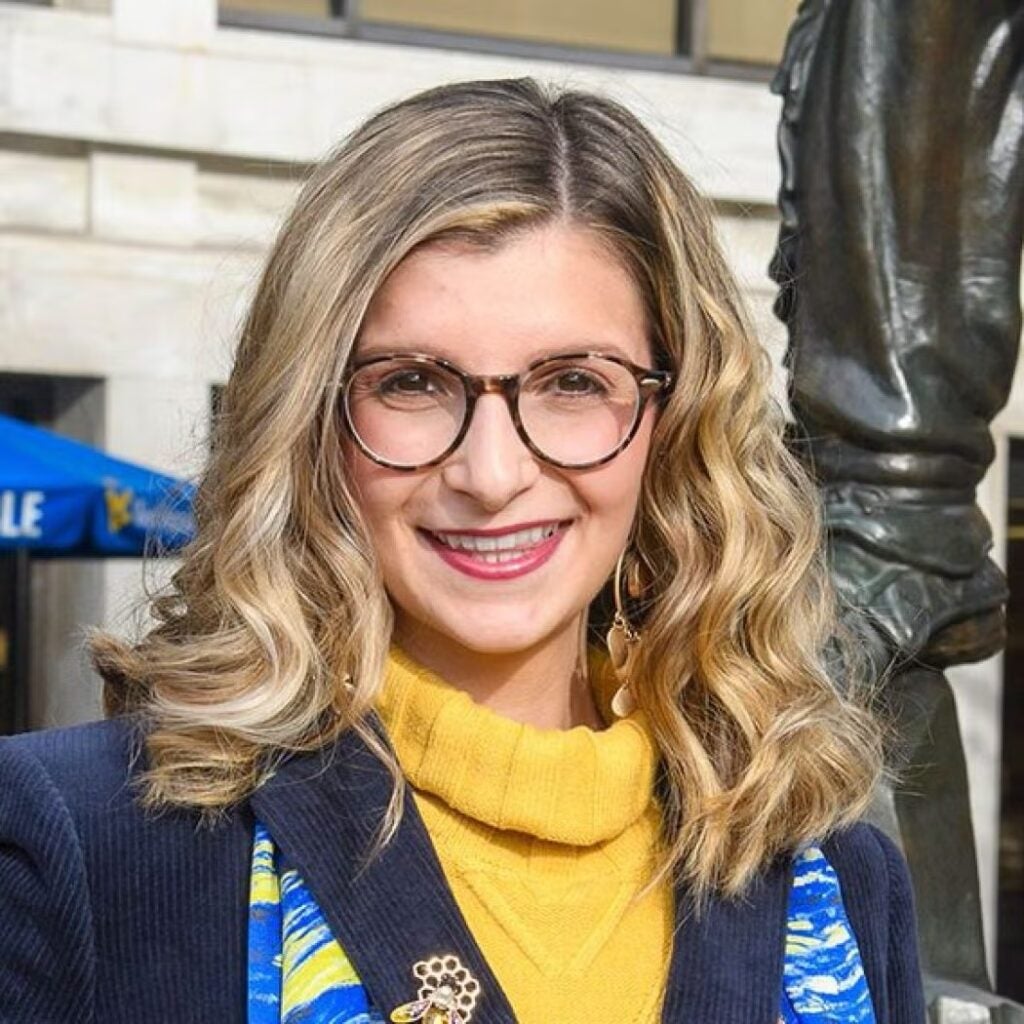 Giana Loretta wearing a navy blue blazer and yellow sweater with glasses.