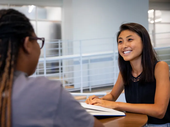 Two women sitting across each other talking and smiling. A book is open on the table that is in the middle of them.