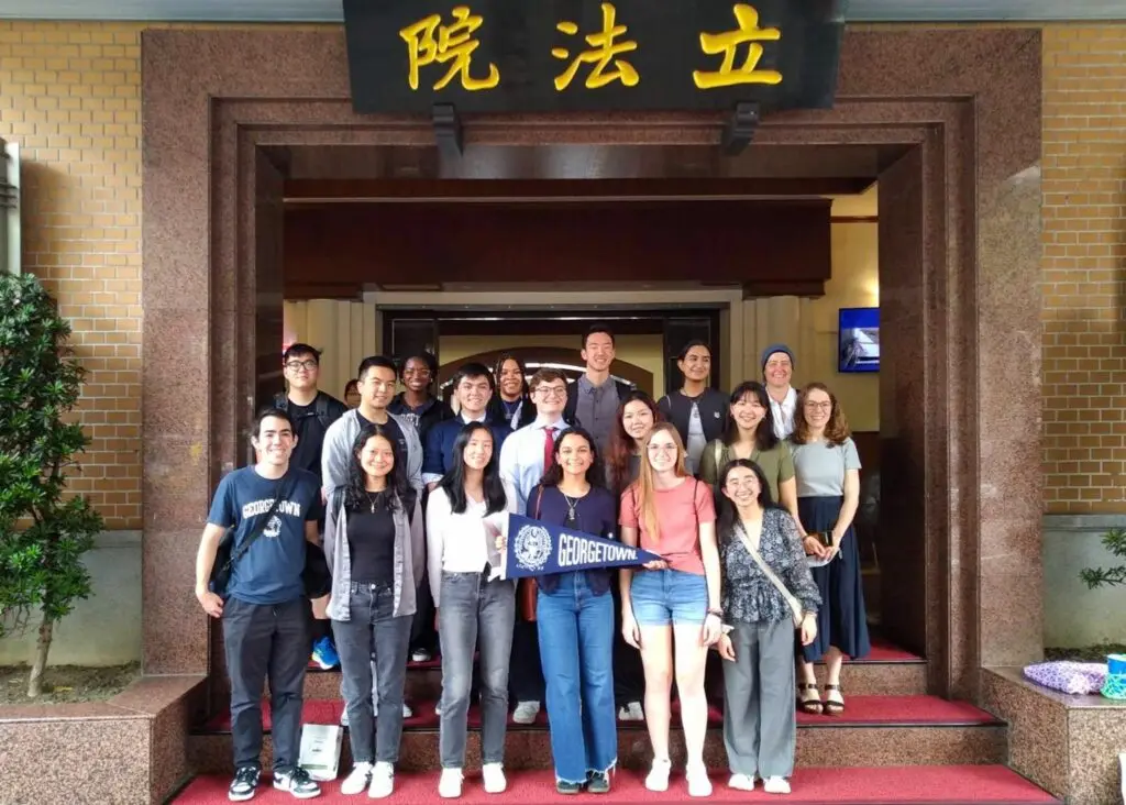 Large group of college students standing on red stairs holding a Georgetown pennant in front of a large square arch opening to a building.