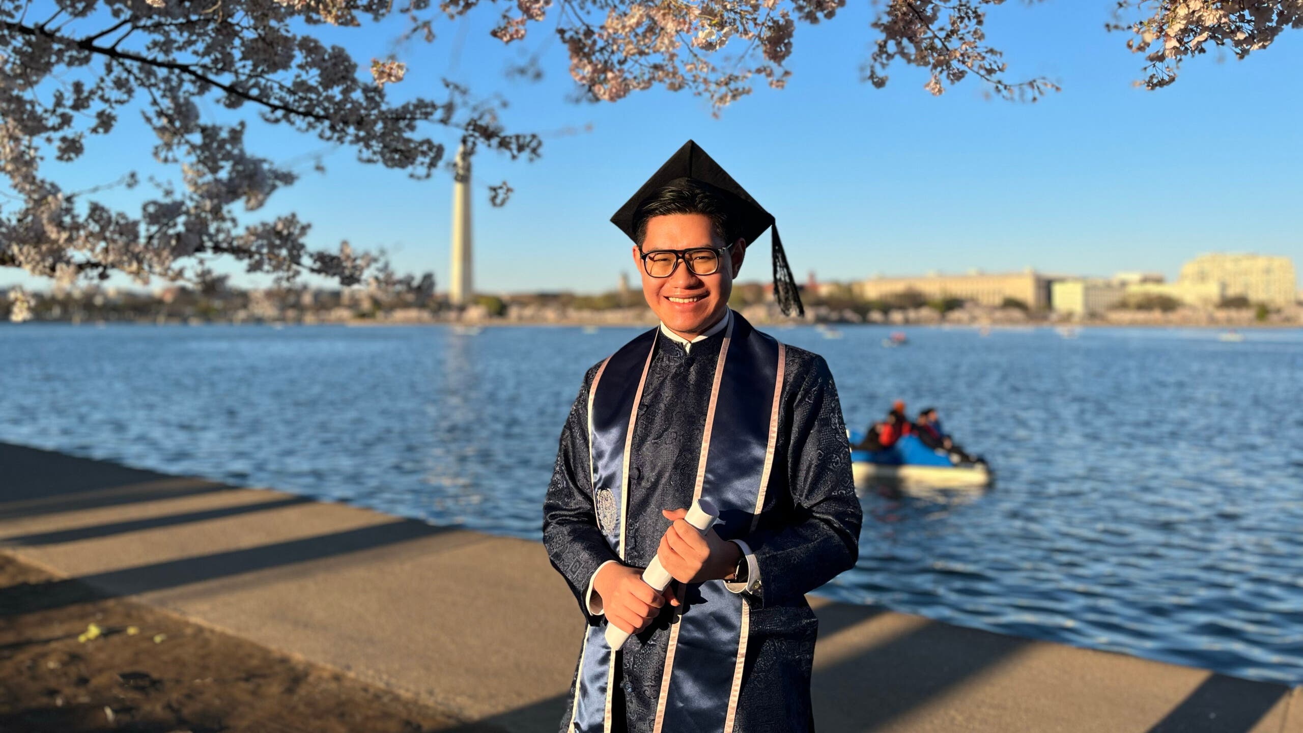 Nguyen stands by the cherry blossoms on the Tidal Basin wearing graduation robes and hat.
