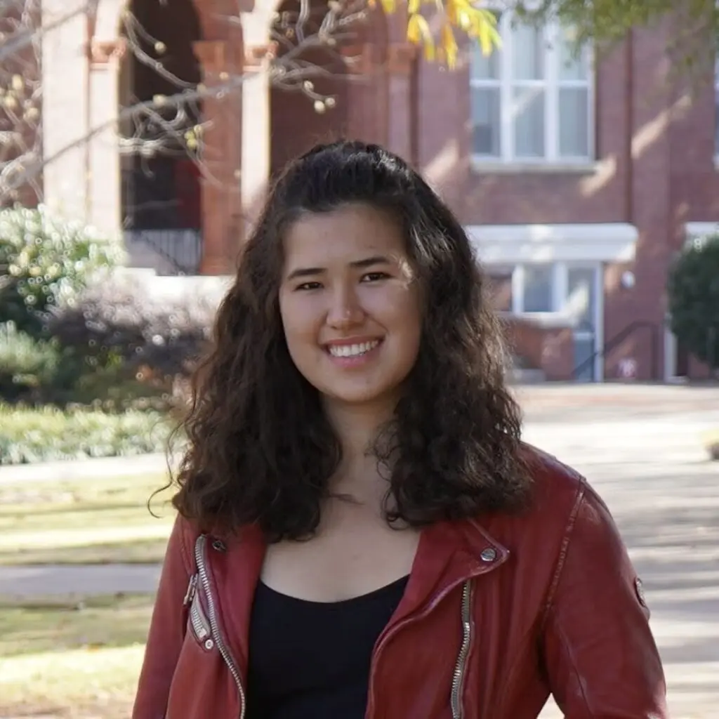 Helga Schroeder wearing a red leather jacket and a black shirt with a brick building in the background.