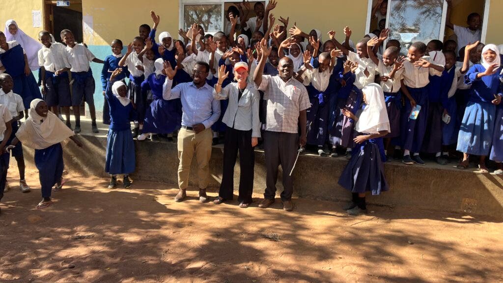 Marina McPhail waves in front of a local elementary school in Tanzania with a group of students and teachers.
