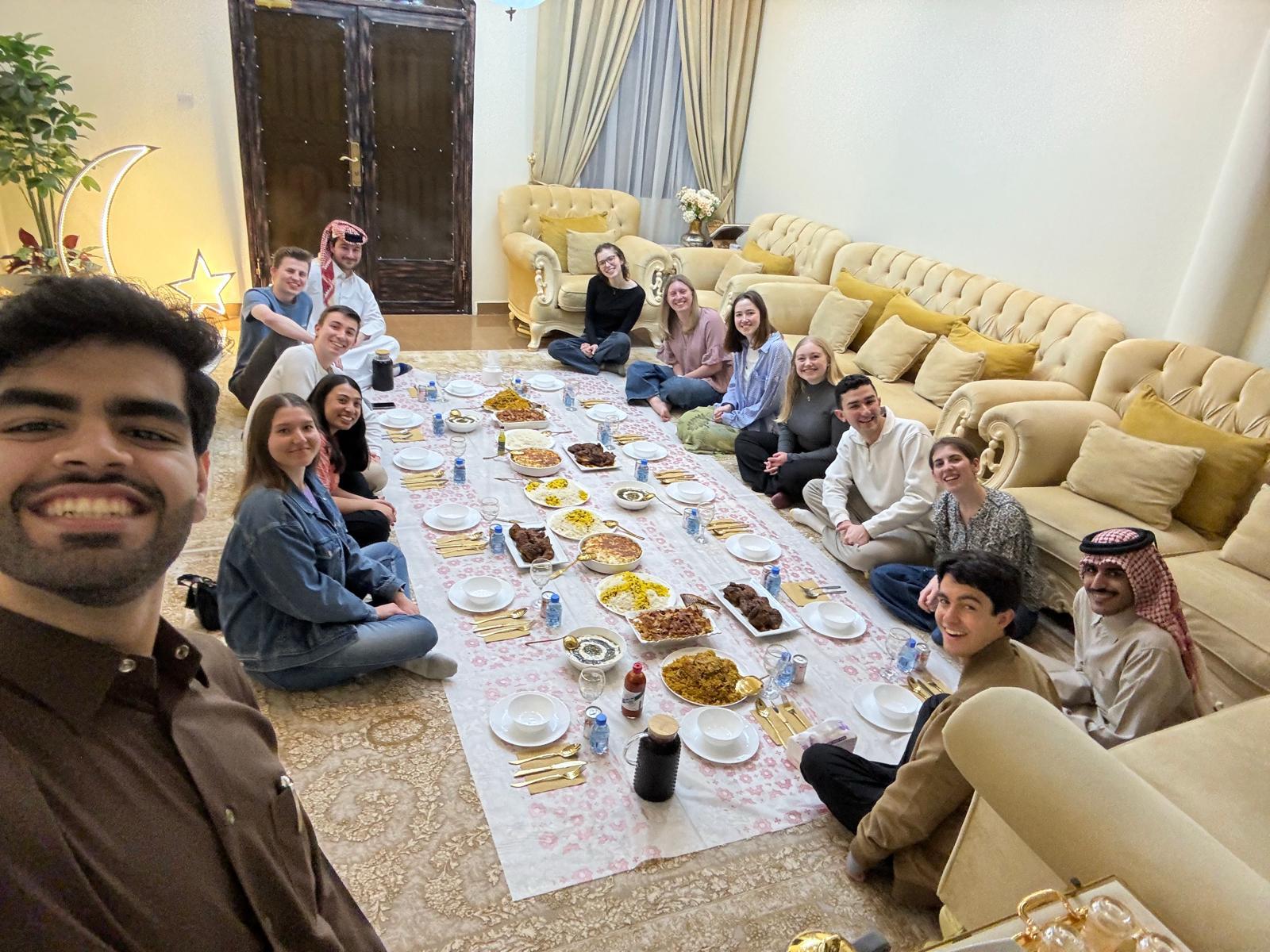 Group of college students sitting around a dining spread on the floor, smiling for a photo in a room with traditional decor.