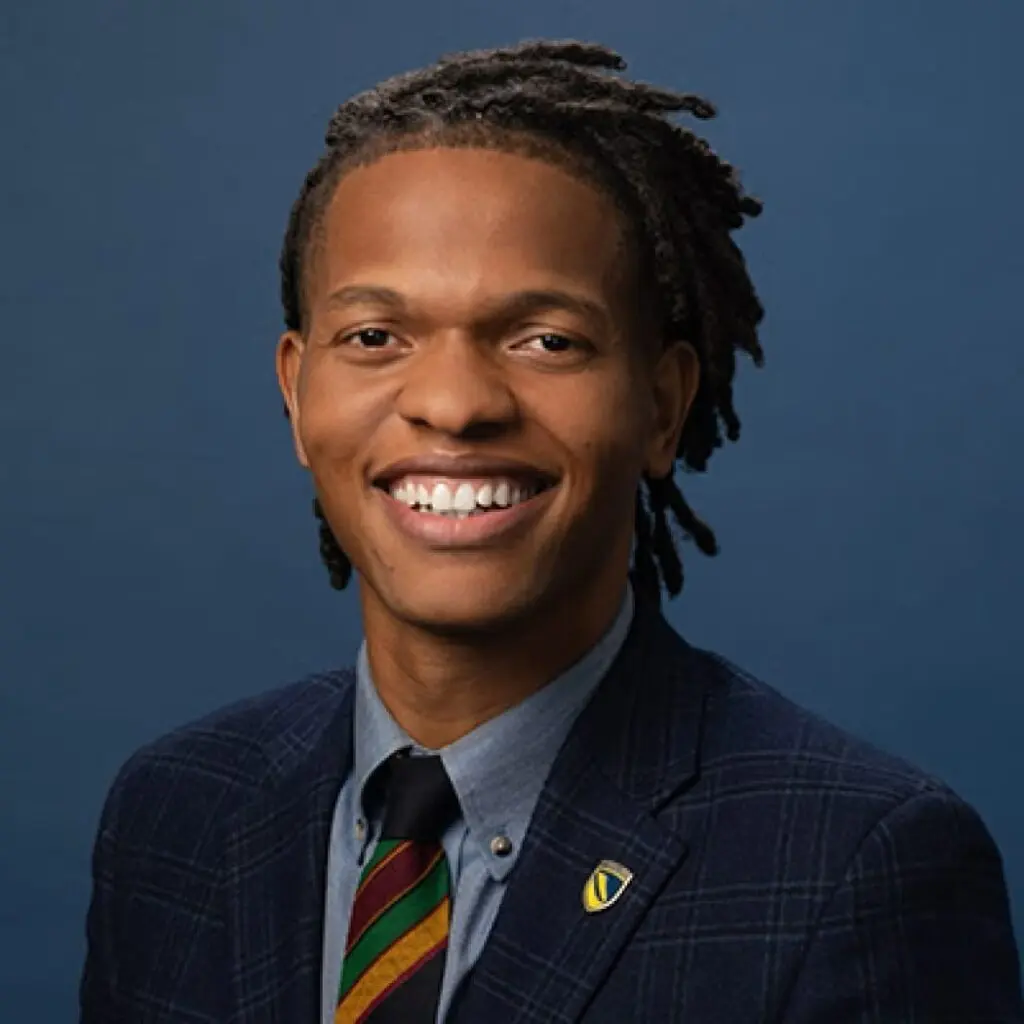 Imhotep Simba wearing a plaid navy blazer, a blue button-down shirt, and multicolored striped tie.