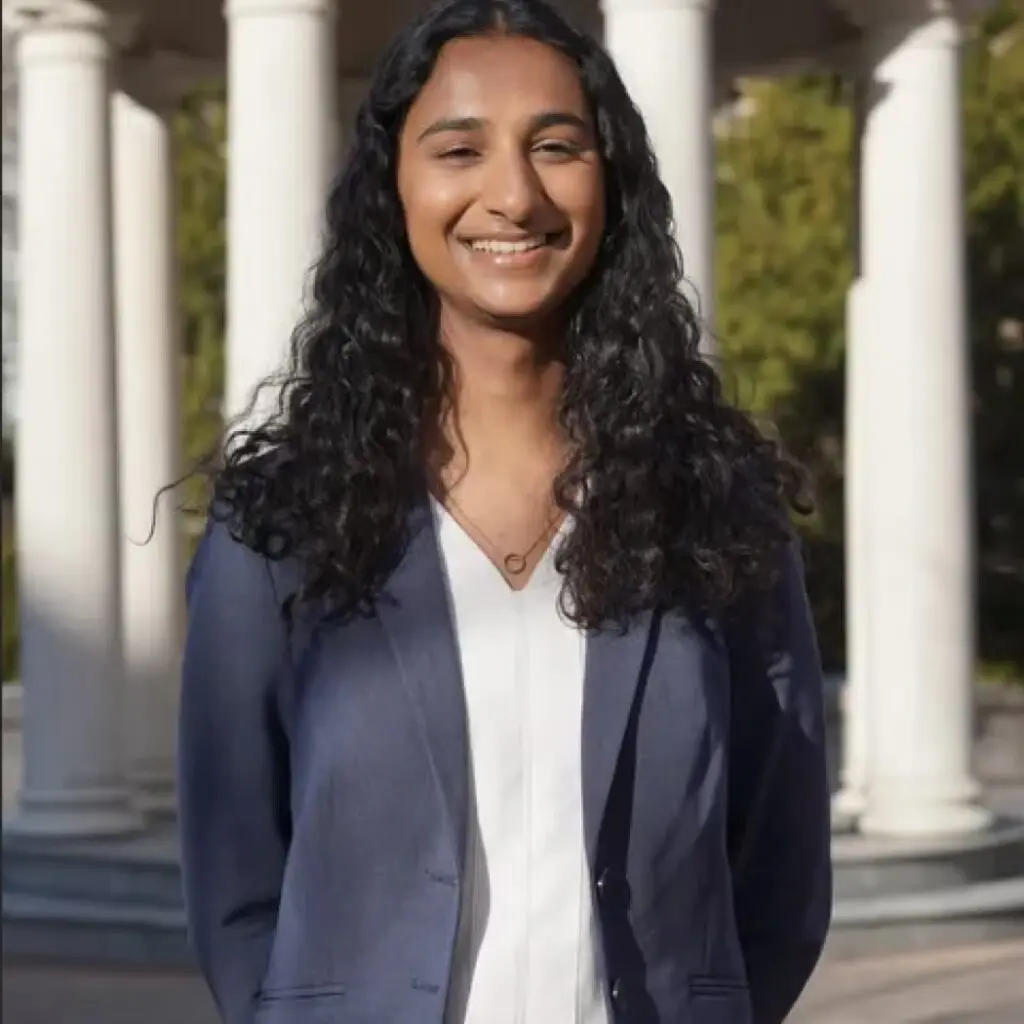 Isha Gupta wearing a navy blue blazer and a white button-down shirt. A building with column is in the background.