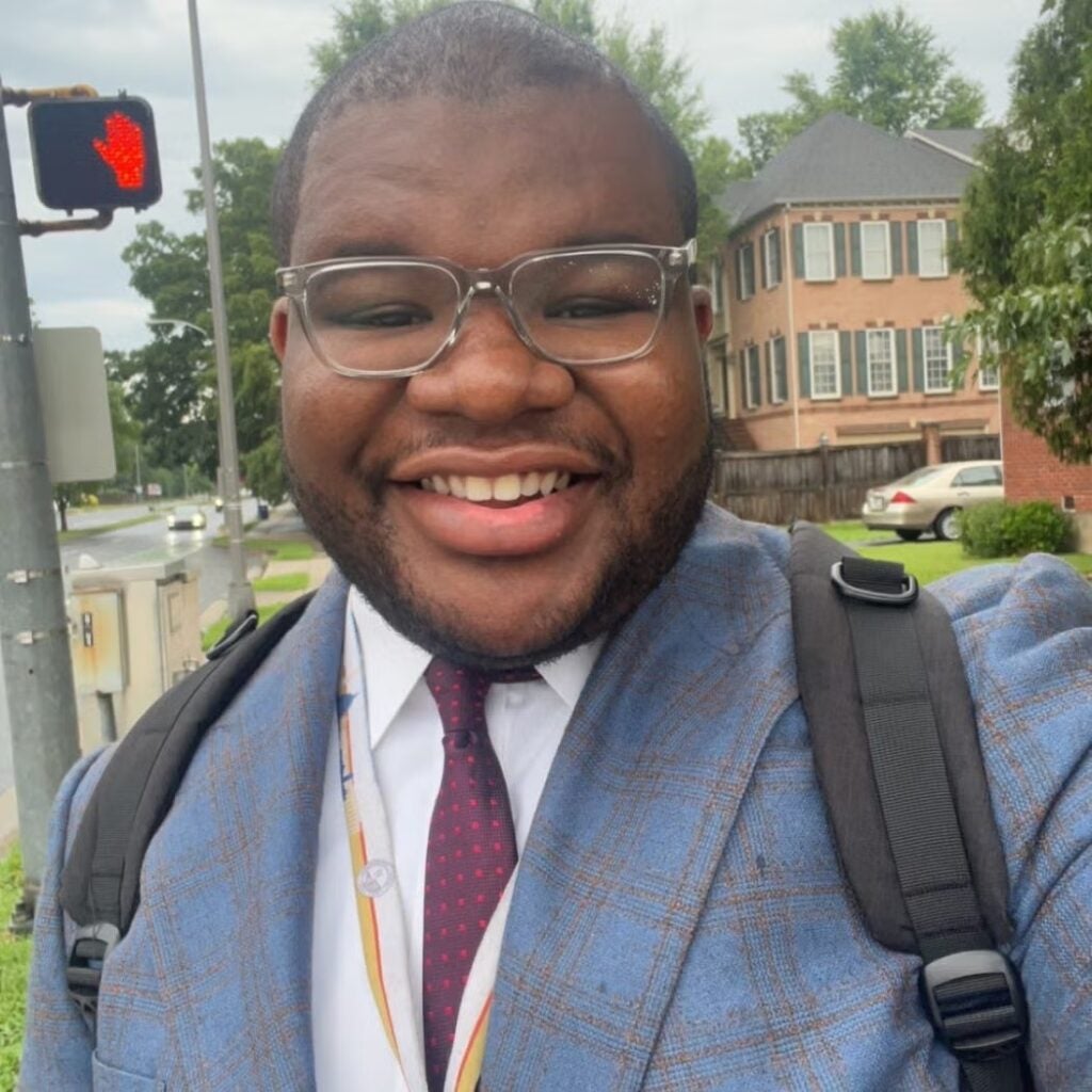 Jamal Halley in a blue and tan plaid blazer with a red tie, white button down shirt carrying a backpack.