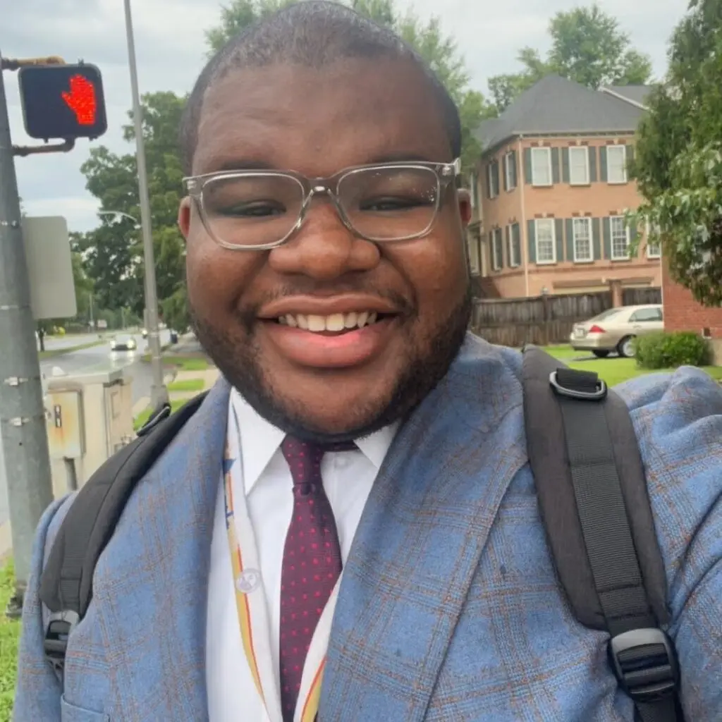 Jamal Halley in a blue and tan plaid blazer with a red tie, white button down shirt carrying a backpack.