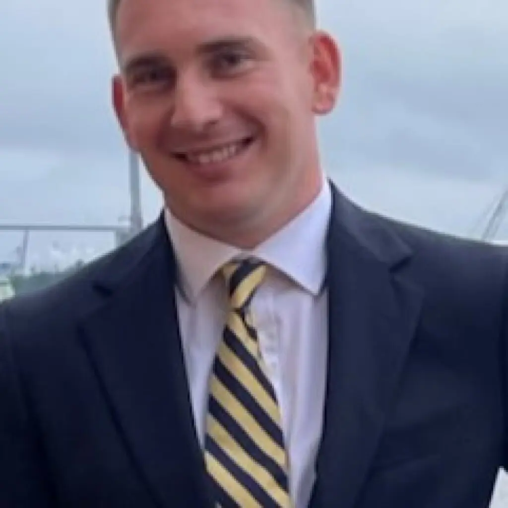 James Bradley wearing a dark blazer and white button-down shirt with a yellow and blue striped tie.