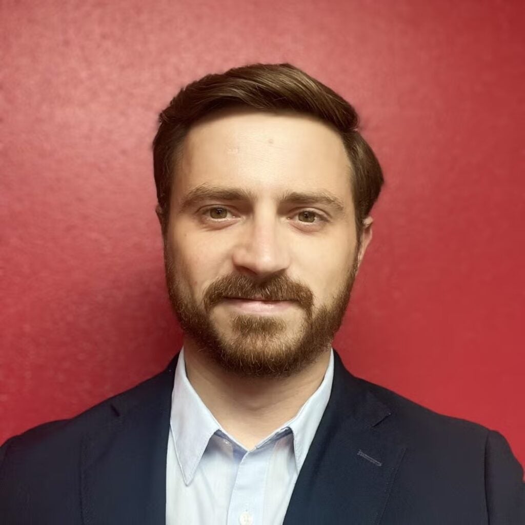 James Olsen wearing a dark blazer and white button-down shirt against a red background.