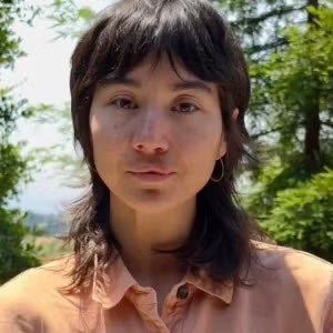 A person with shoulder-length brown hair and a light orange shirt is standing outdoors with trees in the background.