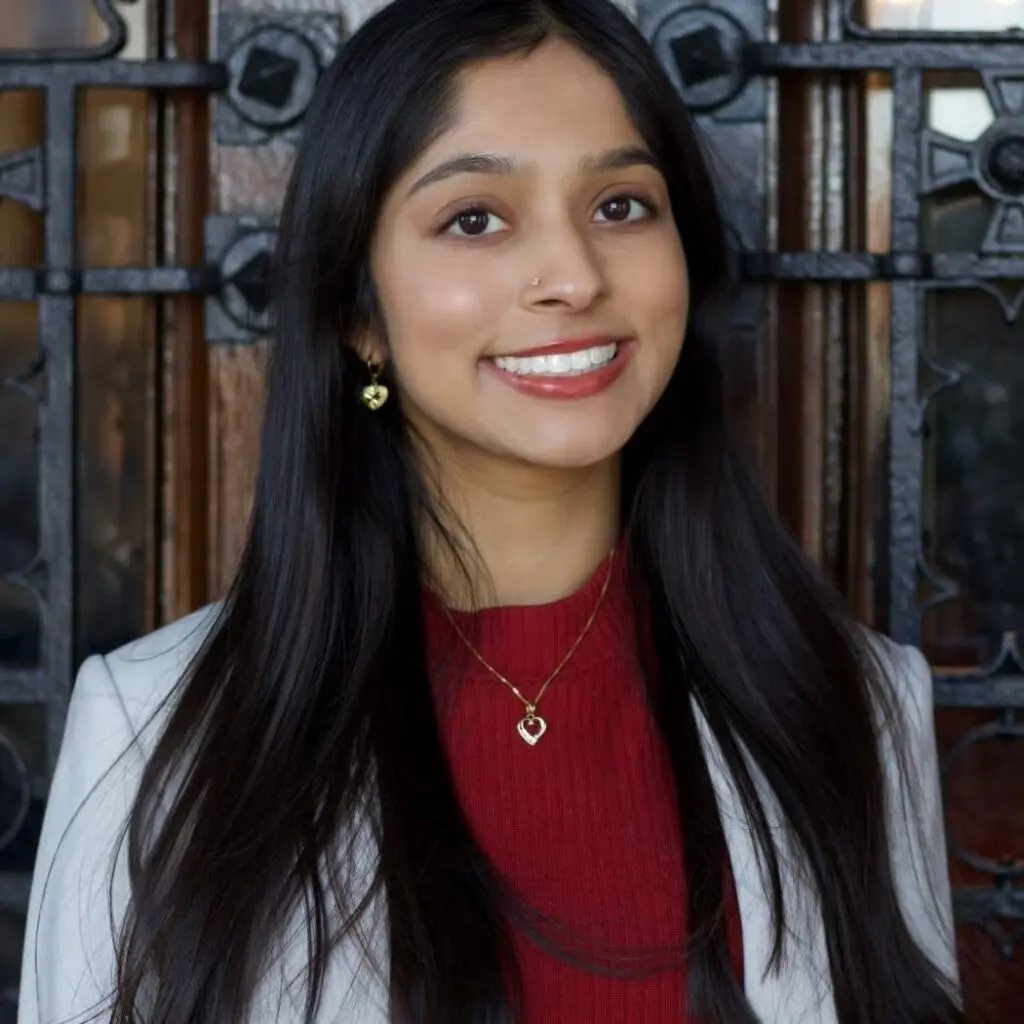 Jasmine Rehman wearing a white blazer with a red sweater and heart shaped necklace and earrings.