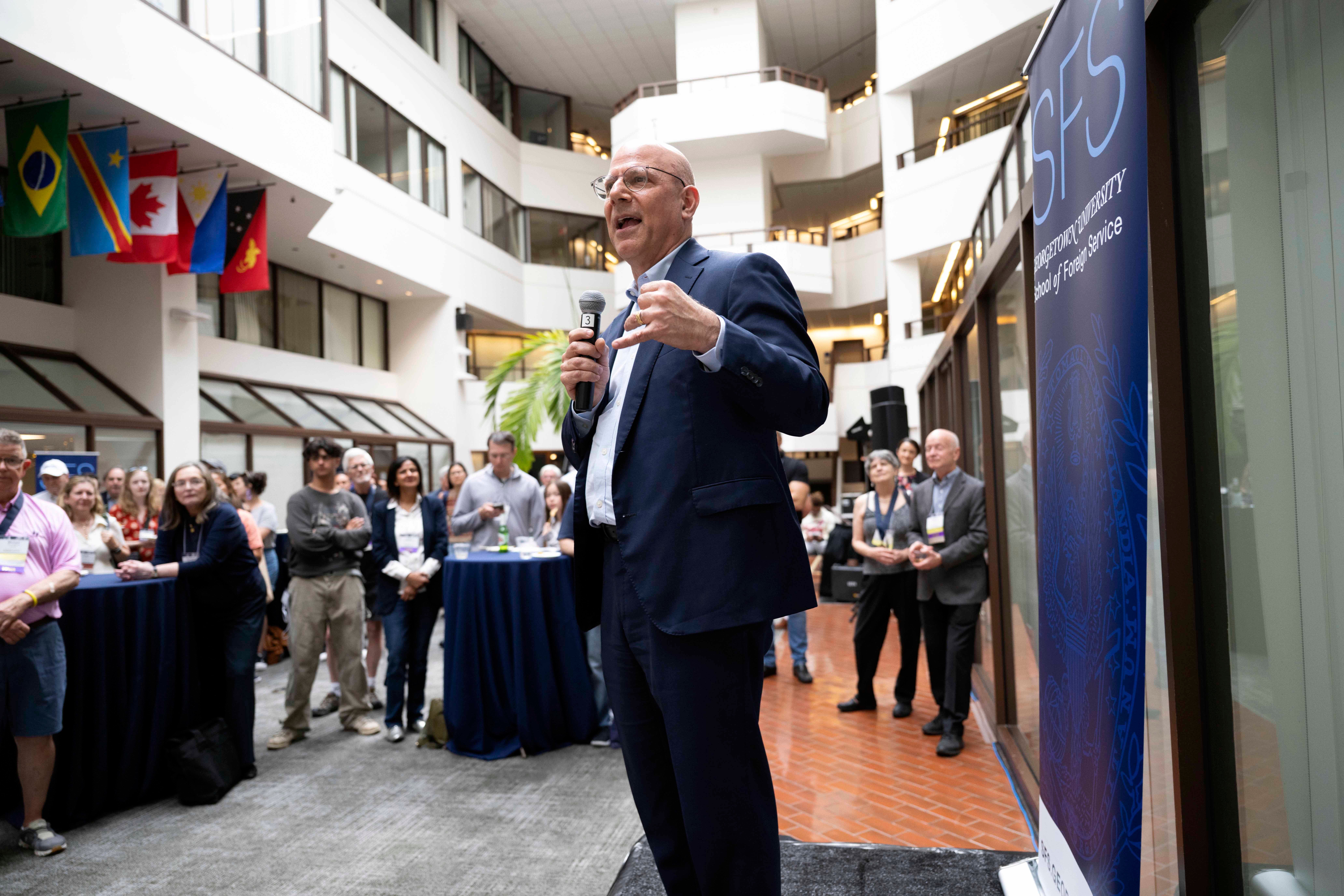 Person delivering a speech at a podium with a microphone in front of an audience in a building with multiple levels and international flags.