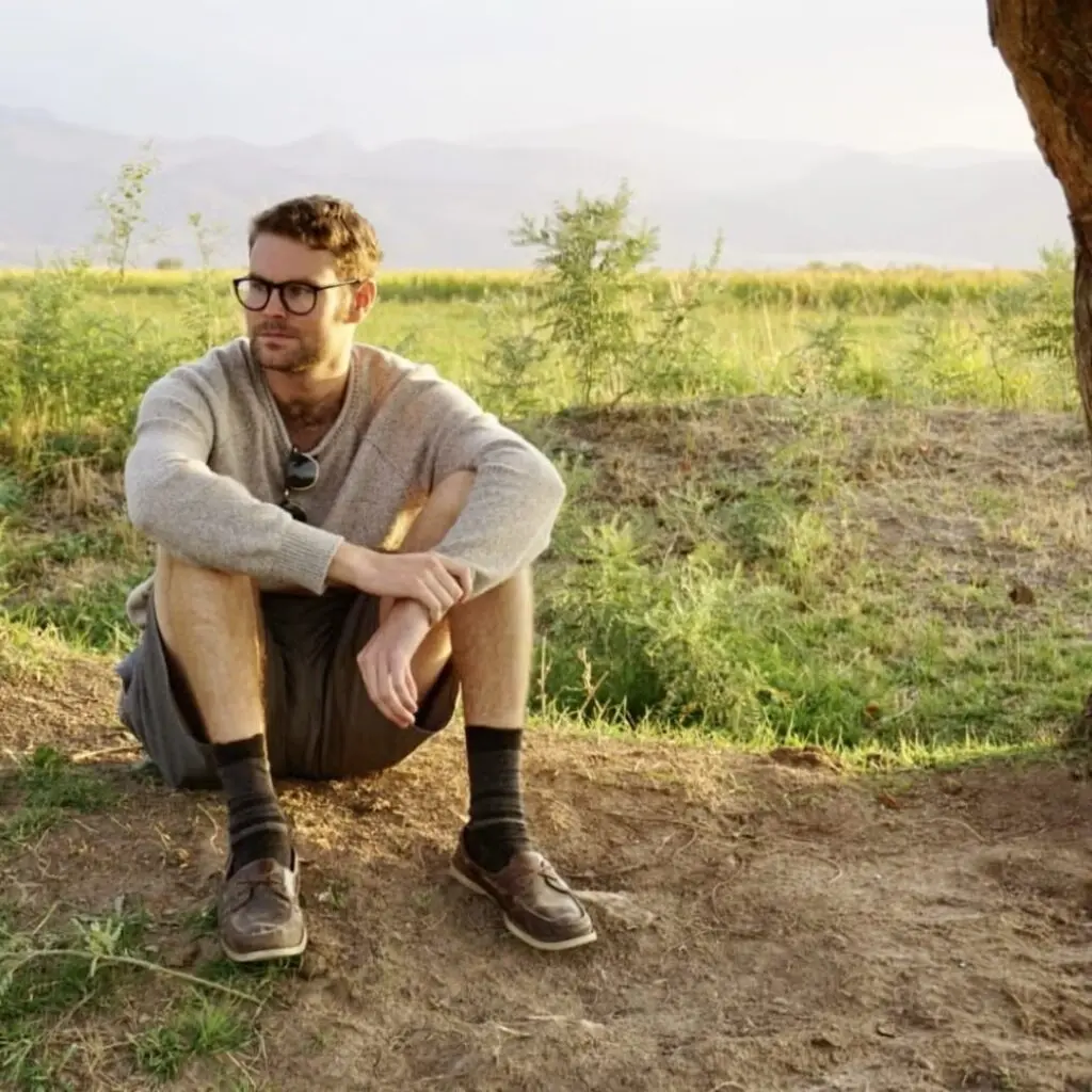 John Murphy sitting in a field, wearing shorts and a tan sweater and wearing glasses.