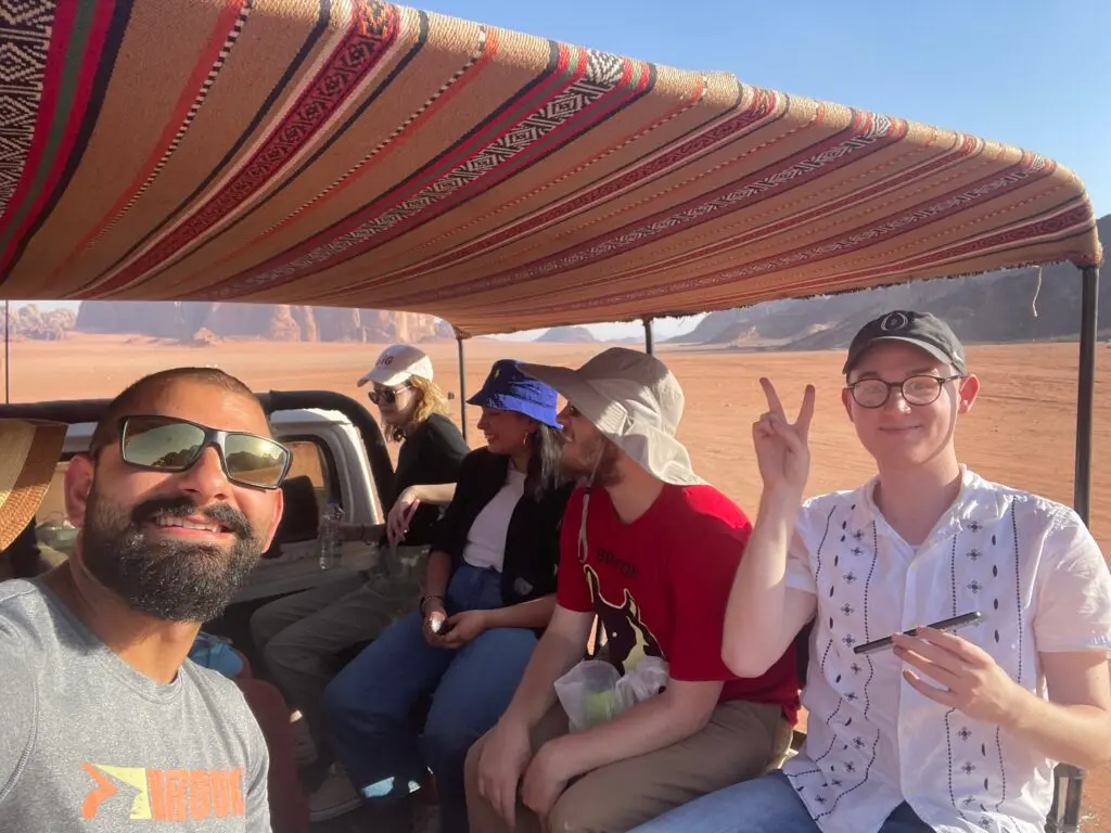 John Korban sits in the back of a truck with a cloth covering with other students with the dessert in the background.