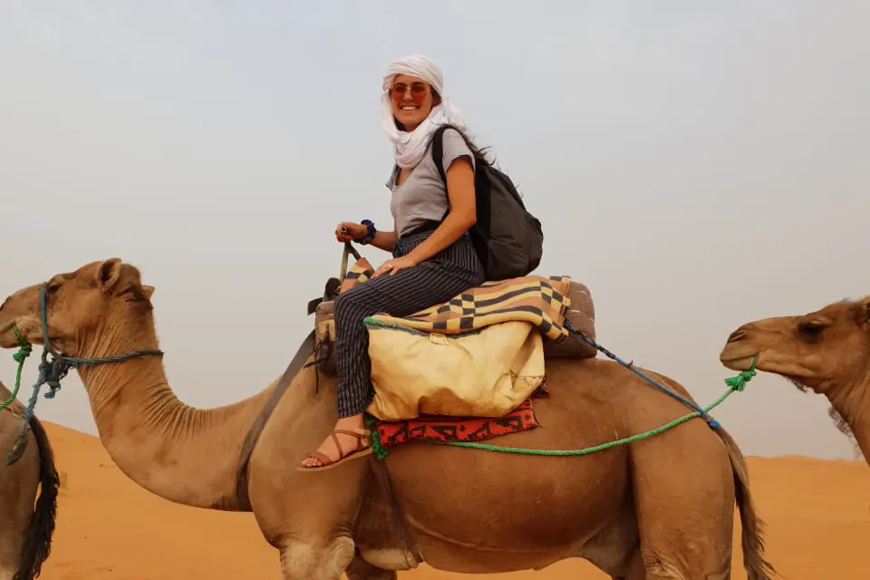 Female student, smiling, riding a camel in the desert over orange sand, and a clear day.