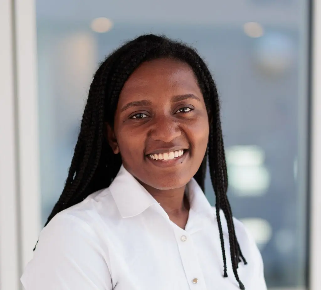 Portrait of a smiling individual with braided hair, wearing a white shirt, standing in front of a blurred office background.