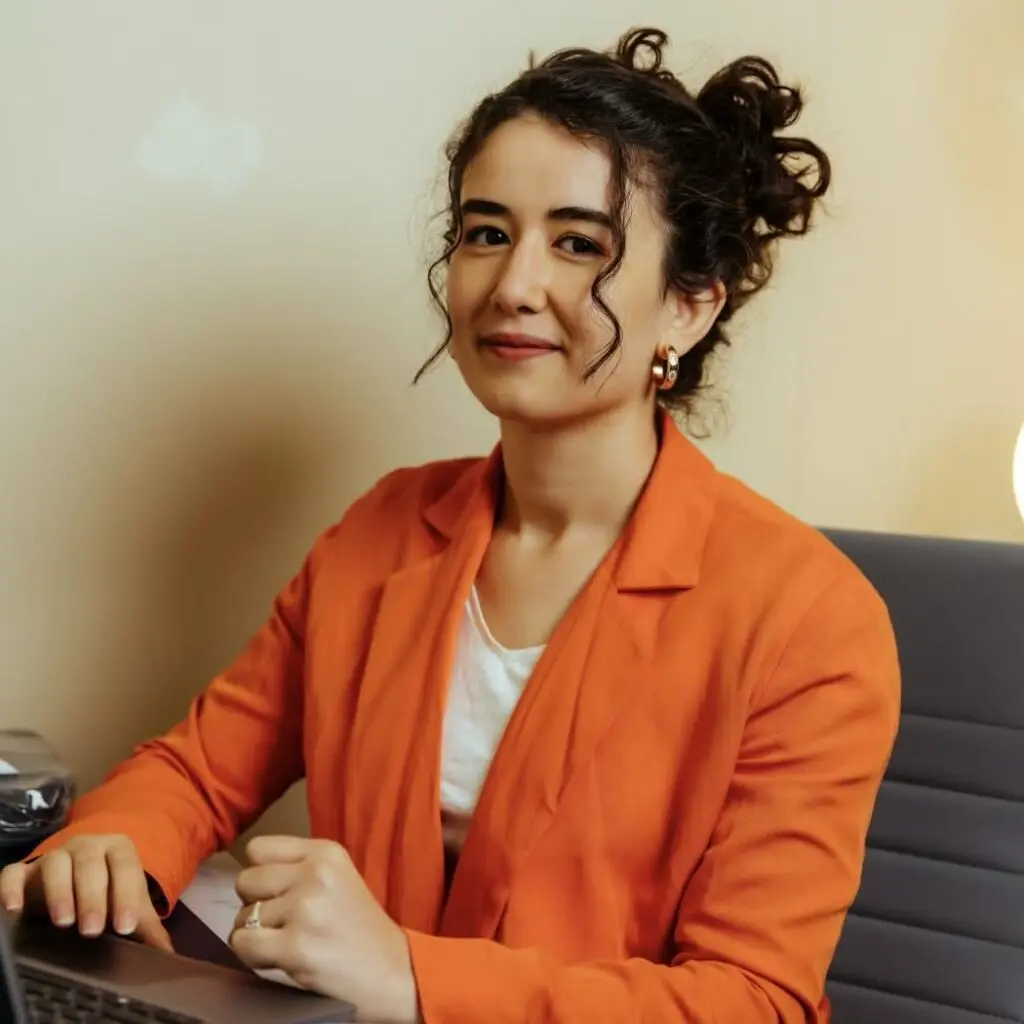 Lamis Ben Aiyad wearing an orange blazer and sitting at a desk.
