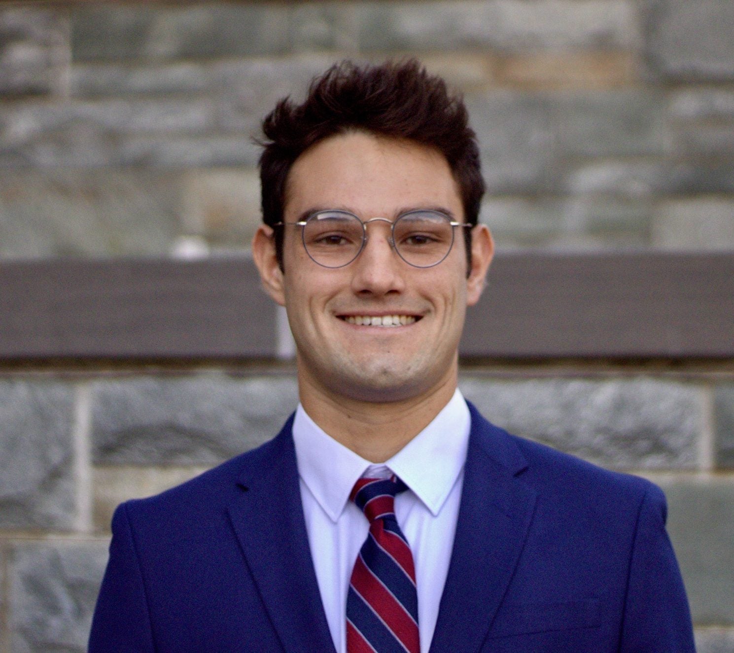 A young man wearing wire-rimmed glasses and a bright blue suit with red and blue striped tie, standing in front of a stone wall.