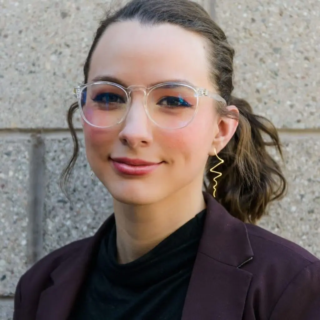 Madisyn GoodBallet wearing a dark purple blazer, a black blouse, gold earrings, and glasses.