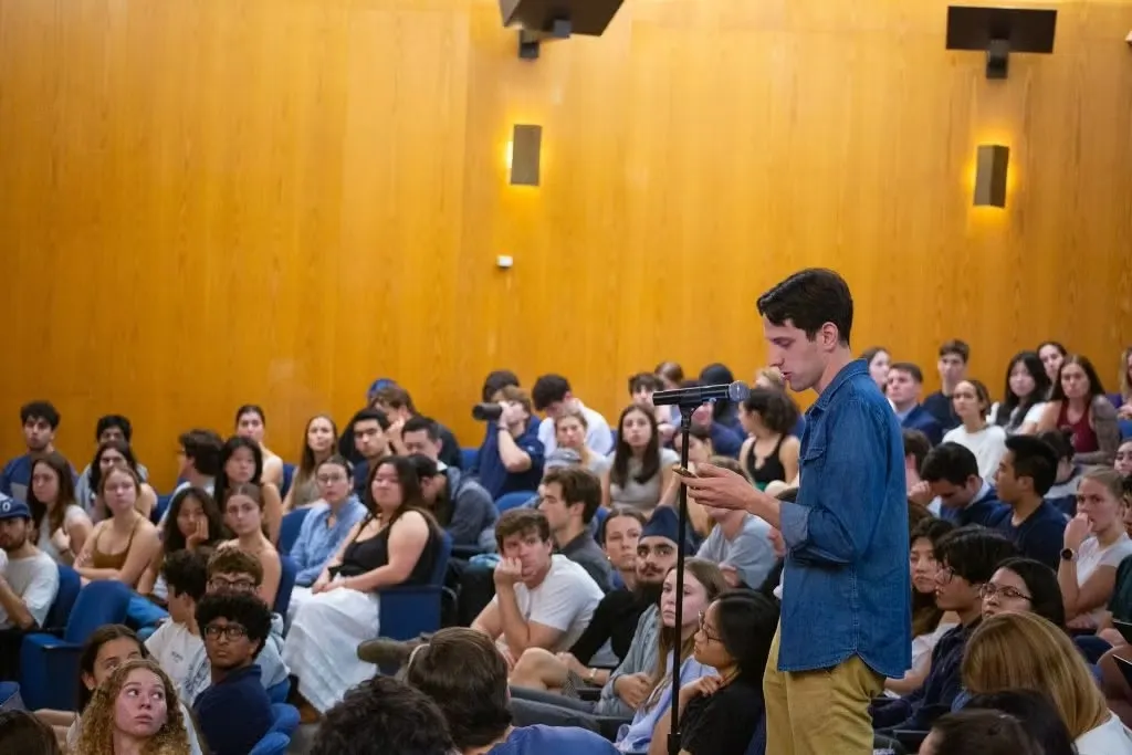 Person wearing a blue shirt standing in front of a microphone reading notes in a crowded auditorium.