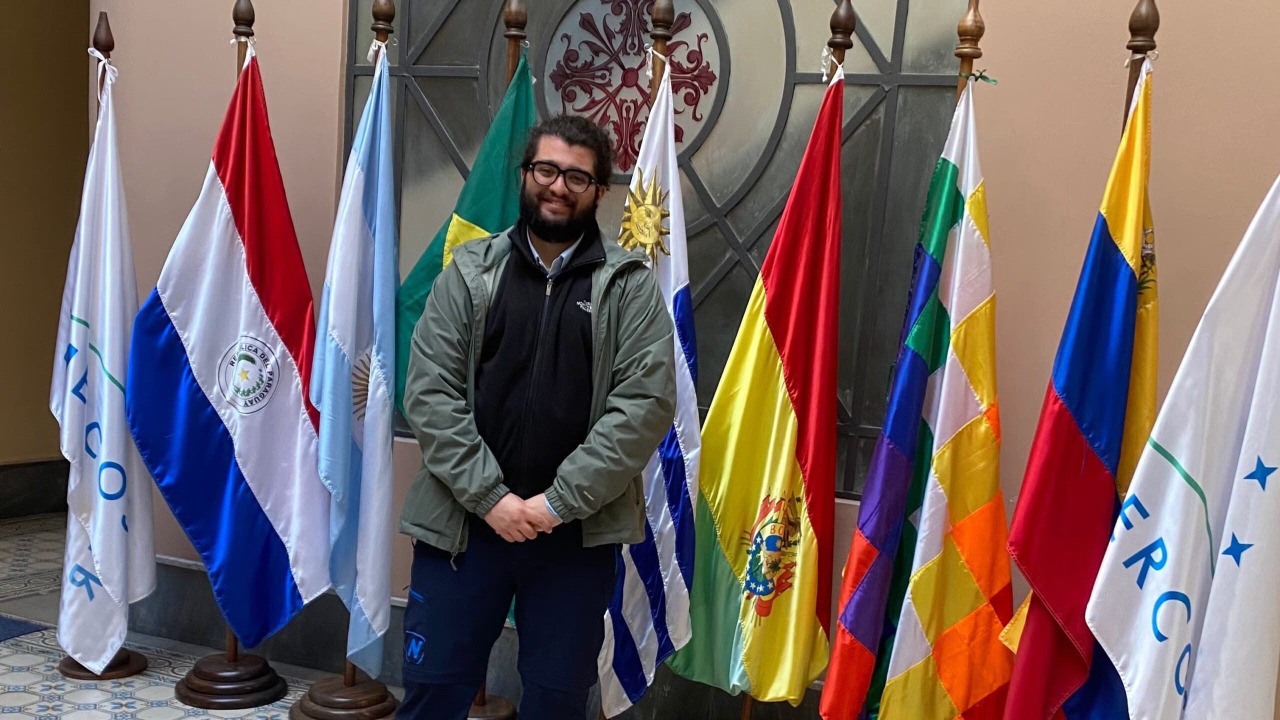 A person smiling and standing in front of a collection of international flags in a tiled hallway.