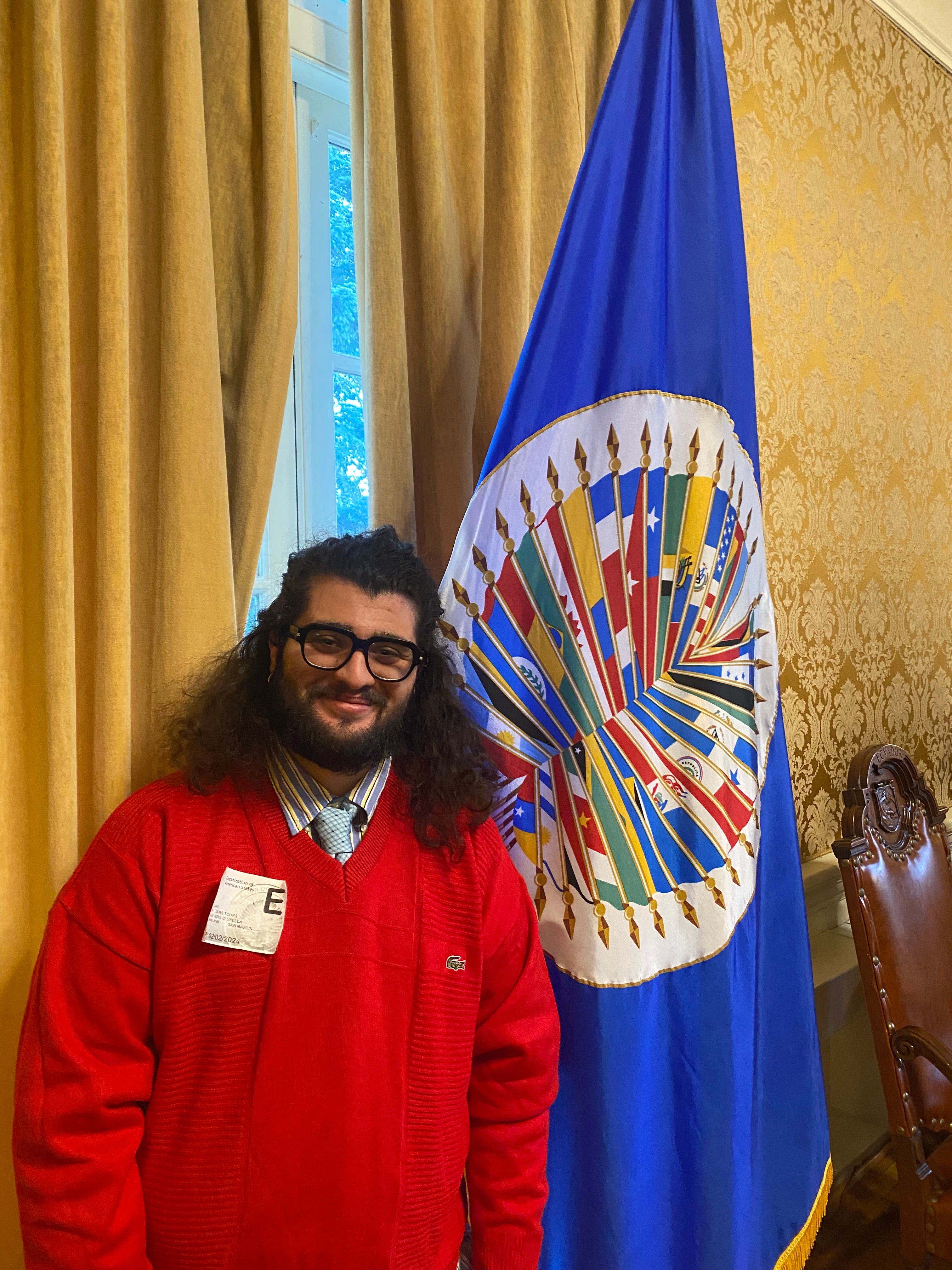 Person wearing a red sweater standing next to the Organization of American States flag in an elegant room.