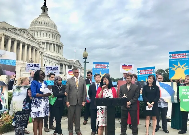 Group of people holding "refugee welcome" signs in front of the US Capitol building. A a woman stand in front of them speaking on a podium.
