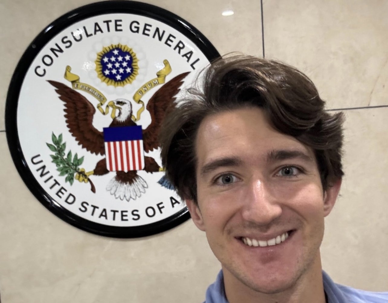 A young man with short brown hair smiling in front of a seal of the Consulate General of the United States.