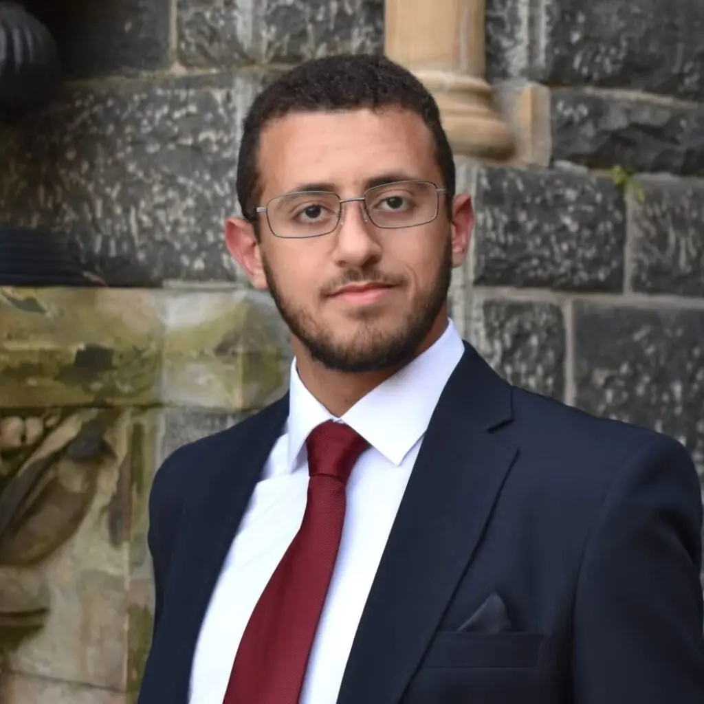 Mohamed Meshal wearing a dark blazer, a burgundy tie, a white shirt and glasses.