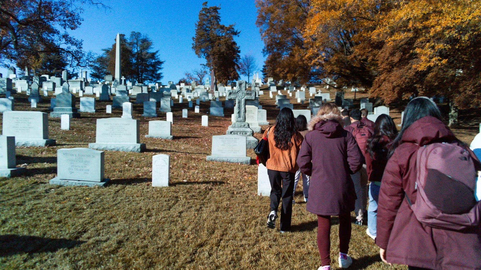 Group of people in winter coats observing gravestones at Arlington National Cemetery, surrounded by autumn-colored trees.