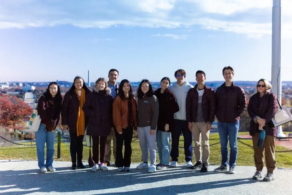 Group of eleven individuals standing together on a sunny day with a clear sky and cityscape in the background.