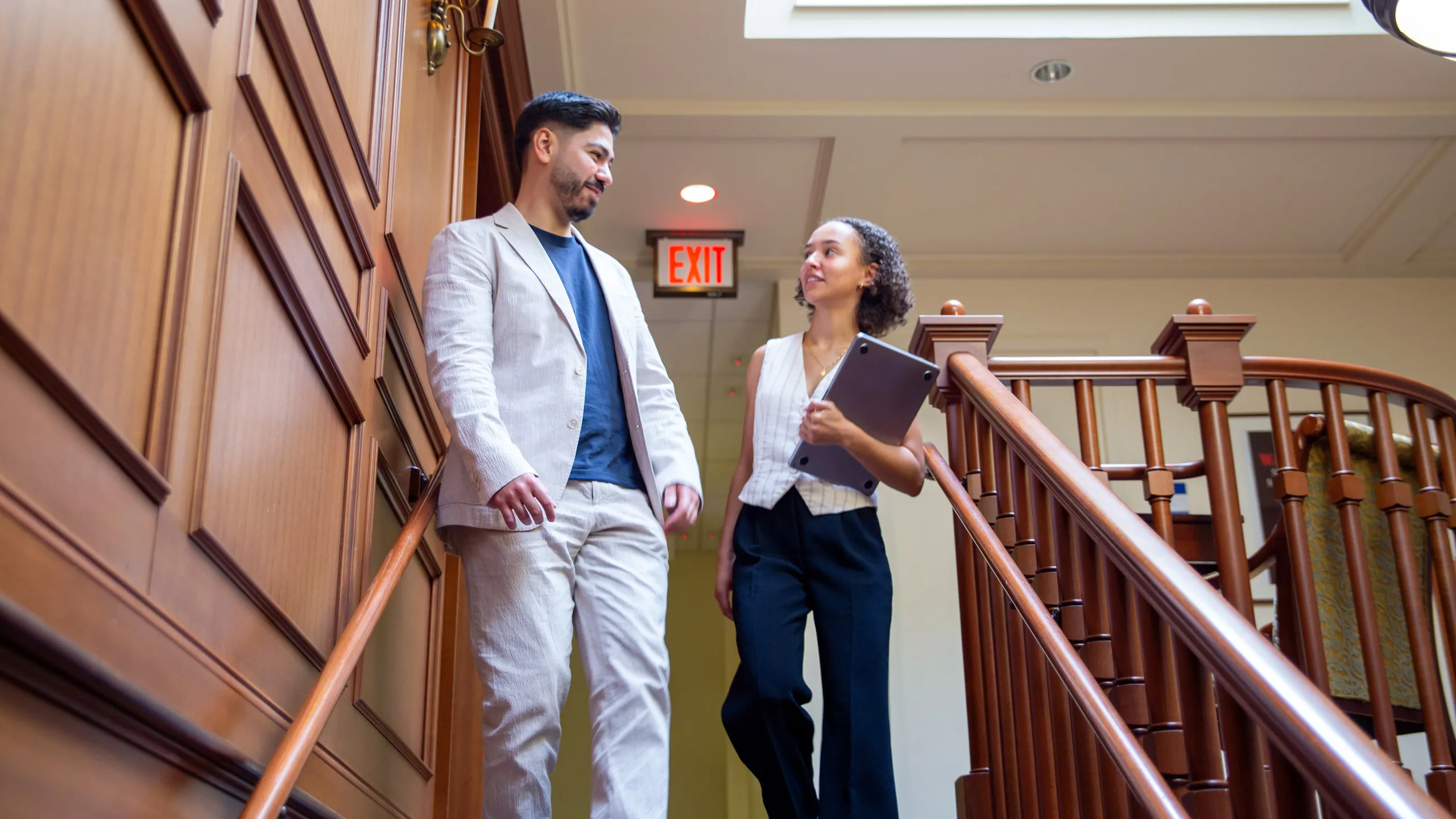 Two students, in business attire, having a conversation while they go down the stairs of a building. The stairs are made out of wood.
