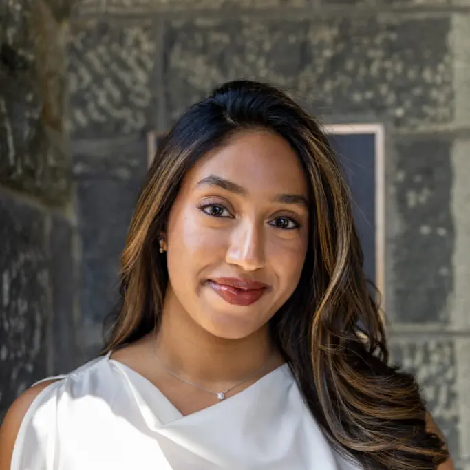 A smiling person with long hair, wearing a white top, standing against a stone wall background.