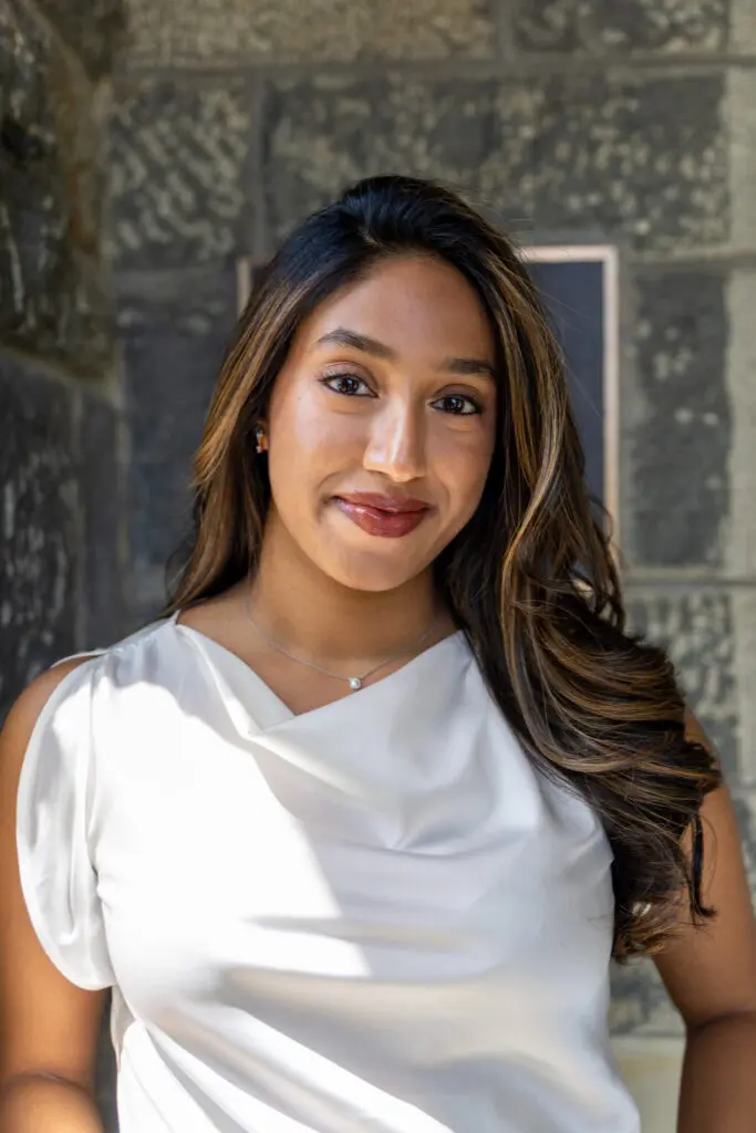 Portrait of a smiling person with long hair, wearing a white top, standing against a stone wall background.