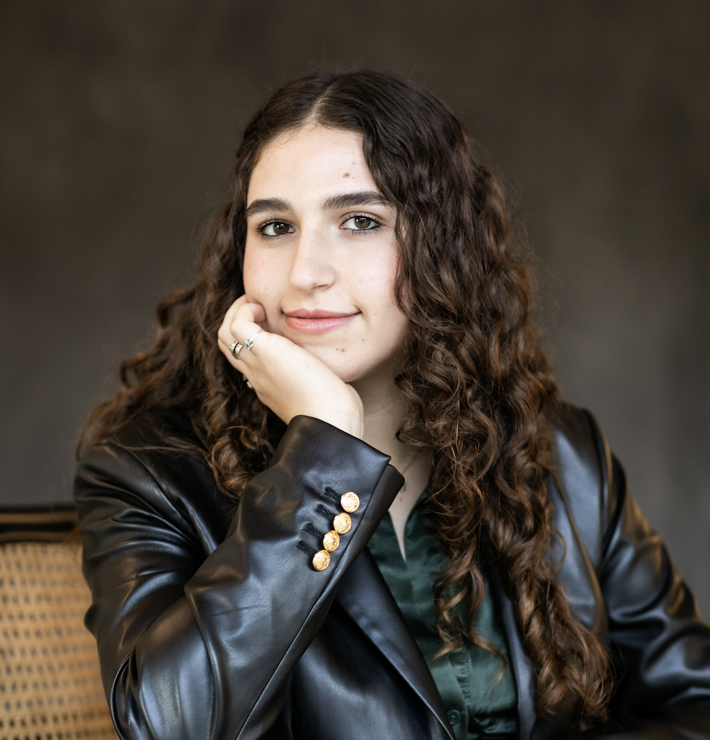 A young woman with long, curly, brown hair wearing a black leather jacket and a dark green shirt.