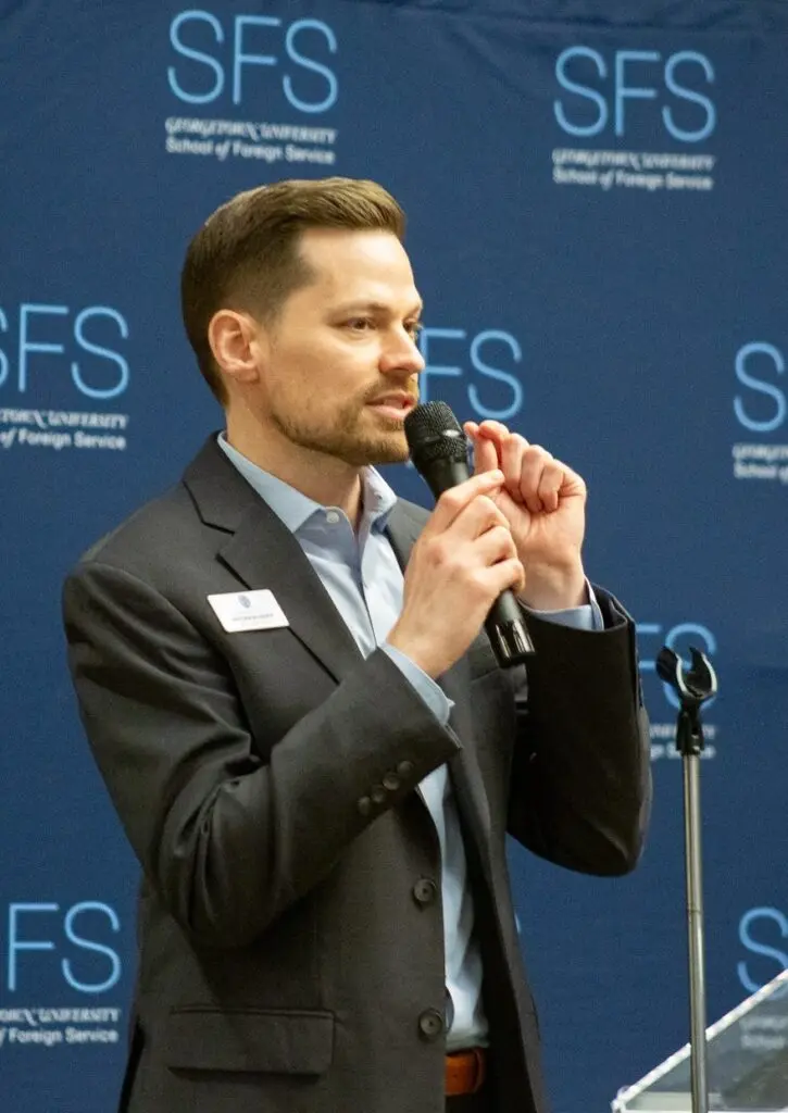 A man in a drak grey suit speaks into a microphone in front of a banner that says "SFS."