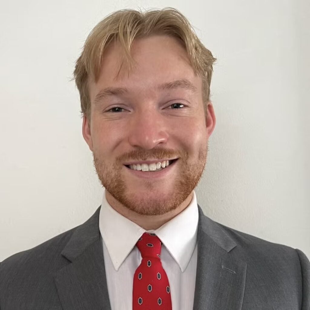 Nicholas McCurrach wearing a gray blazer with a white button-down shirt and a red tie.