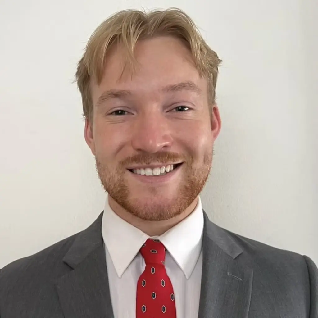 Nicholas McCurrach wearing a gray blazer with a white button-down shirt and a red tie.