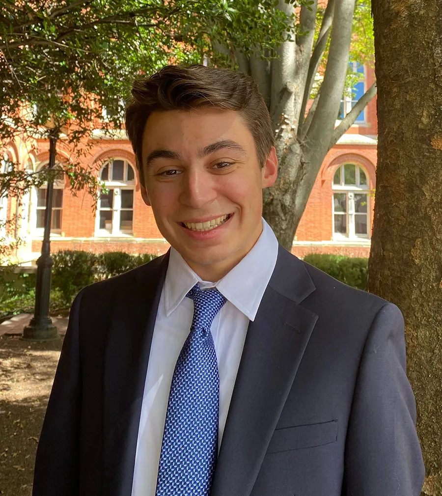A young man with short brown hair wearing a dark blue suite and a blue tie, smiling in front of a tree and a brick building.