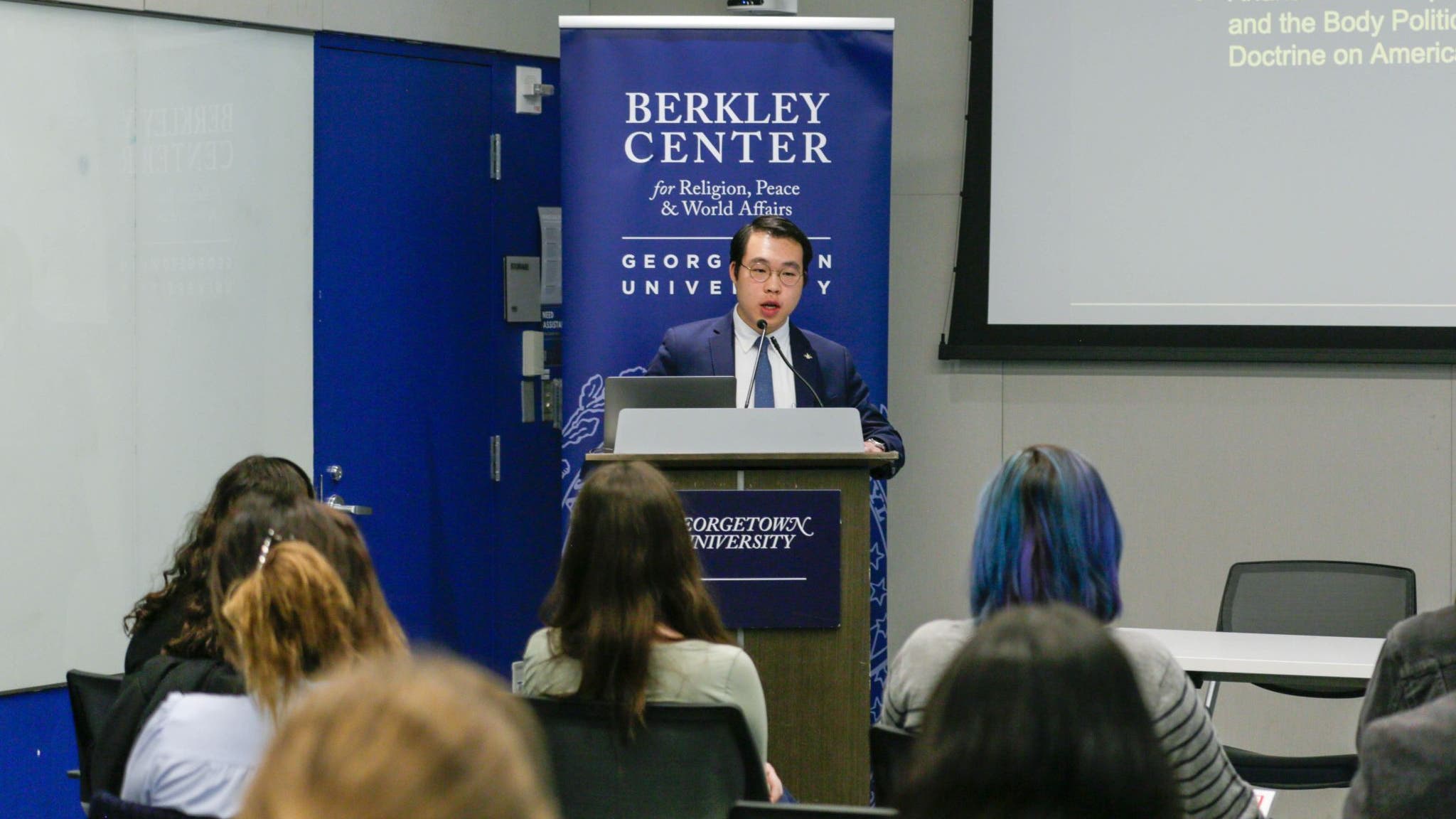 An individual speaks at a podium, in a room with attendees and banners labeled "Berkley Center for Religion, Peace & World Affairs.