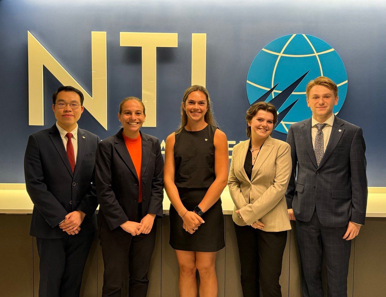 Five individuals smiling for a photo in front of a wall featuring the logo of NTI. They are dressed in professional business attire.