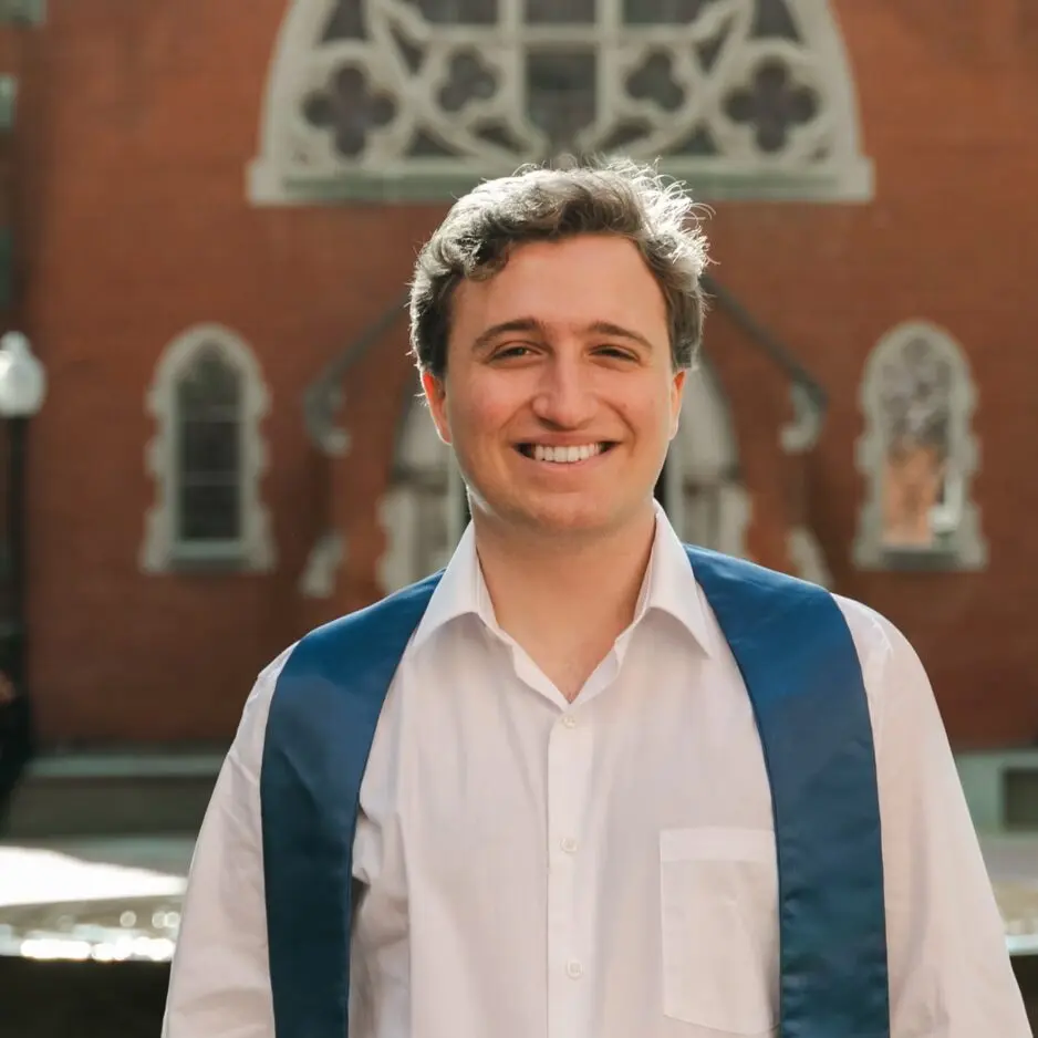Headshot of a smiling Ben Oestericher, wearing a white shirt, he stands in front of a brick building.