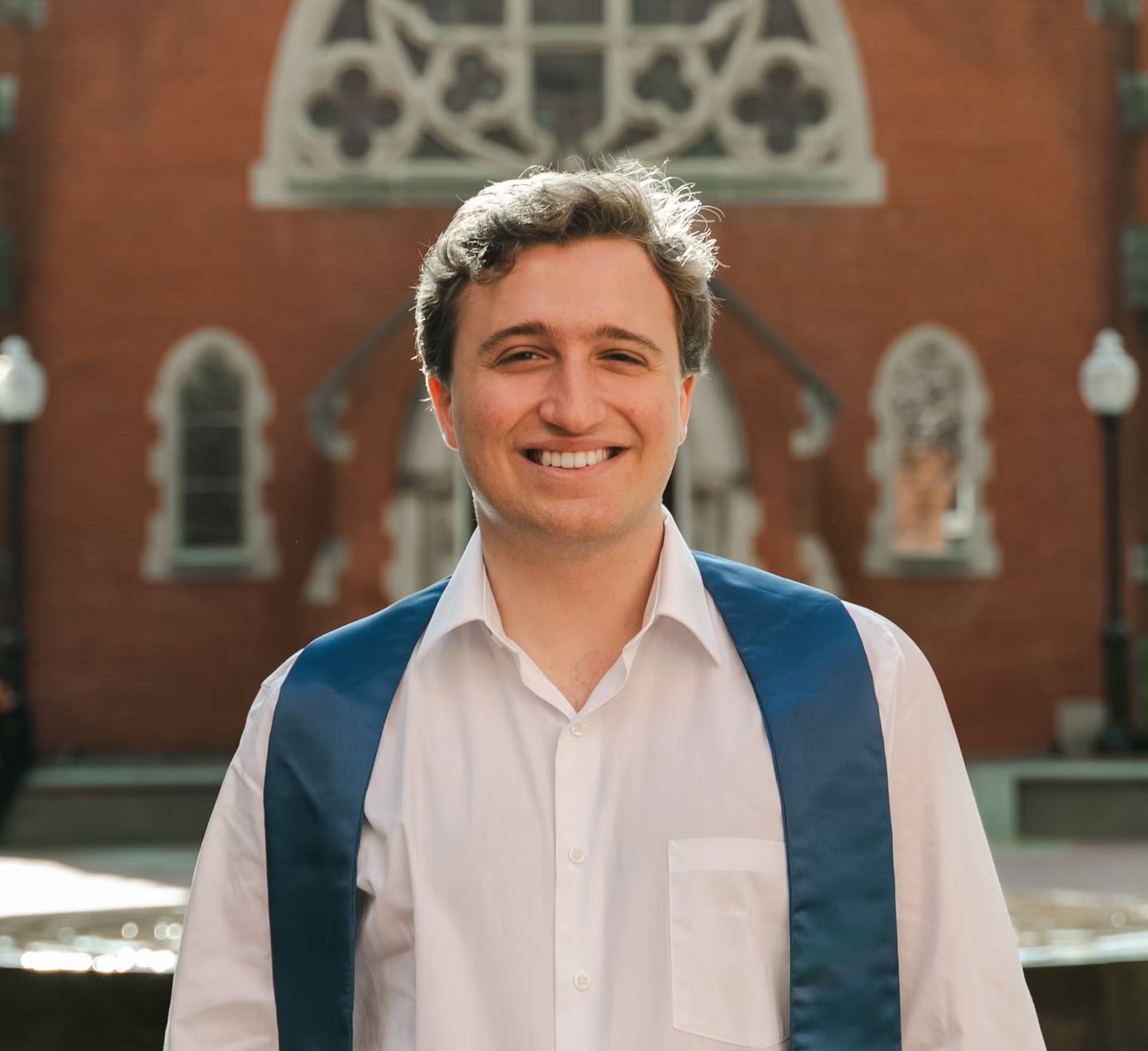 Headshot of a smiling Ben Oestericher, wearing a white shirt in front of a brick building.