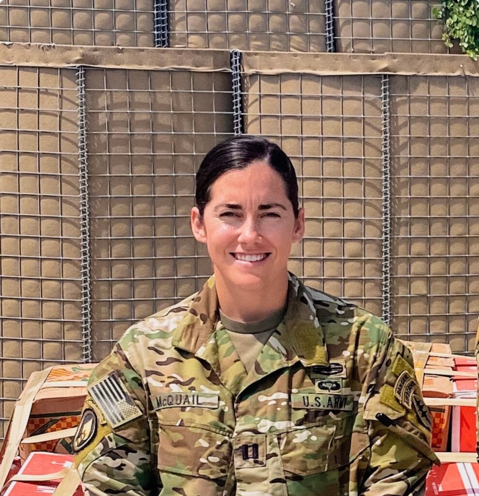 Person in U.S. Army uniform smiling, with a sandbag barrier in the background.