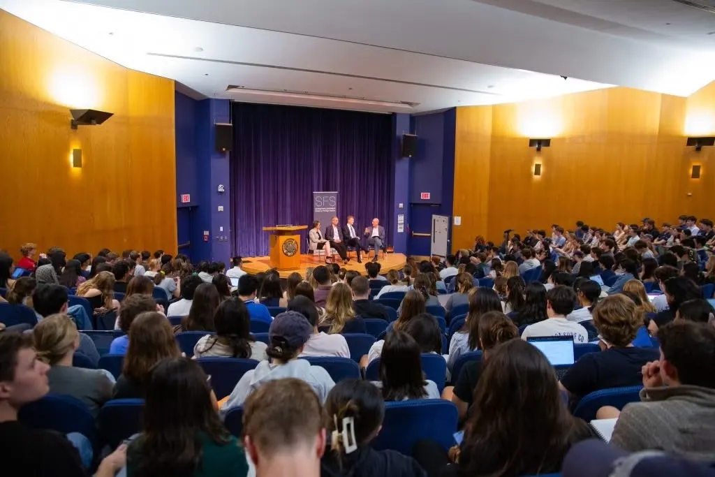 Audience of students at a lecture in an auditorium at the School of Foreign Service, with three speakers seated on stage under a banner reading "SFS."