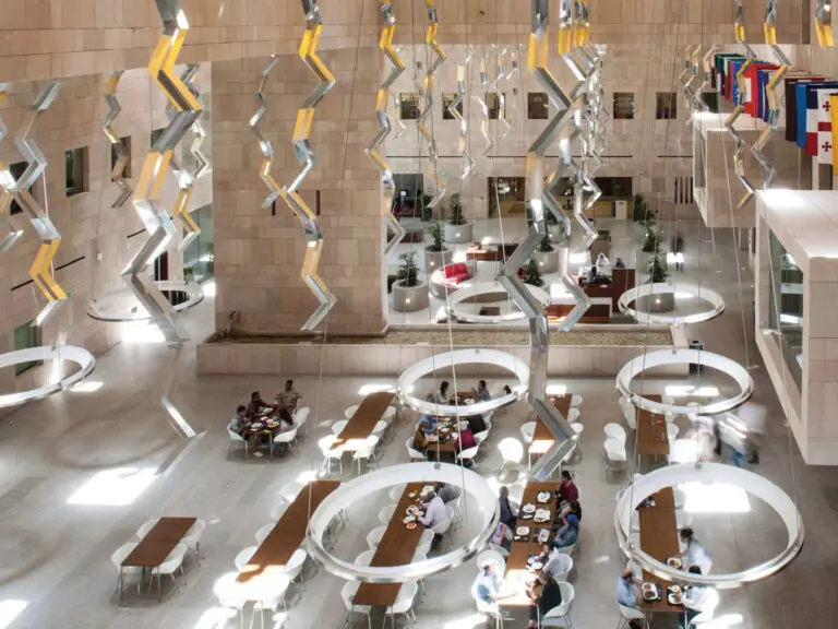 Inside of a light colored spacious building. Flags hang on the right side wall. Tables at the bottom floor where people engage in conversations.